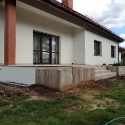 Nowoczesny dom jednorodzinny z tarasem w trakcie budowy, widoczne betonowe elementy konstrukcyjne tarasu, świeżo obsypane ziemią, w tle budynki mieszkalne i zieleń.