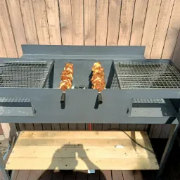 Szary, metalowy grill z dwoma ruszami i drewnianą półką, na którym znajdują się dwa szaszłyki w trakcie grillowania, na tle drewnianej ściany.