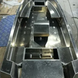 Wnętrze aluminiowej łodzi motorowej w trakcie budowy, widoczne spawane łączenia i surowe wykończenie metalu, siedziska i schowki z pokrywami, widok z góry.