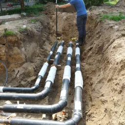 Geodeta mierzy poziom wykopu z rurami, prawdopodobnie pod instalację geotermalną, używając niwelatora w otoczeniu ziemi i zieleni.