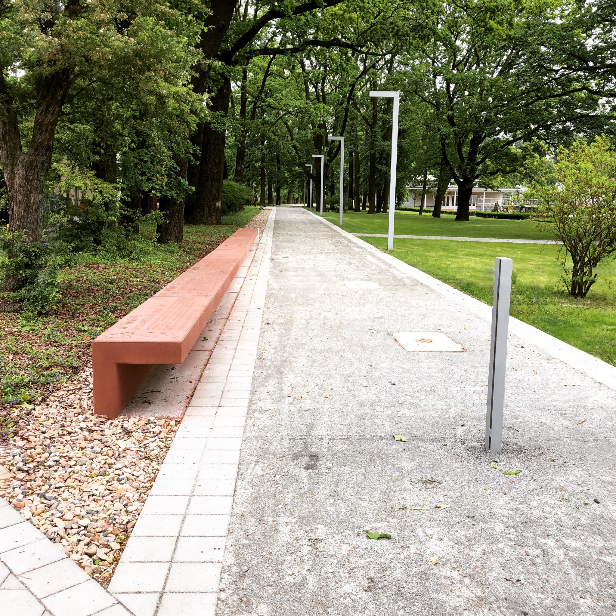 Betonowa ławka z inskrypcjami wzdłuż parkowej ścieżki oświetlonej nowoczesnymi lampami, otoczona zielenią drzew i trawników.