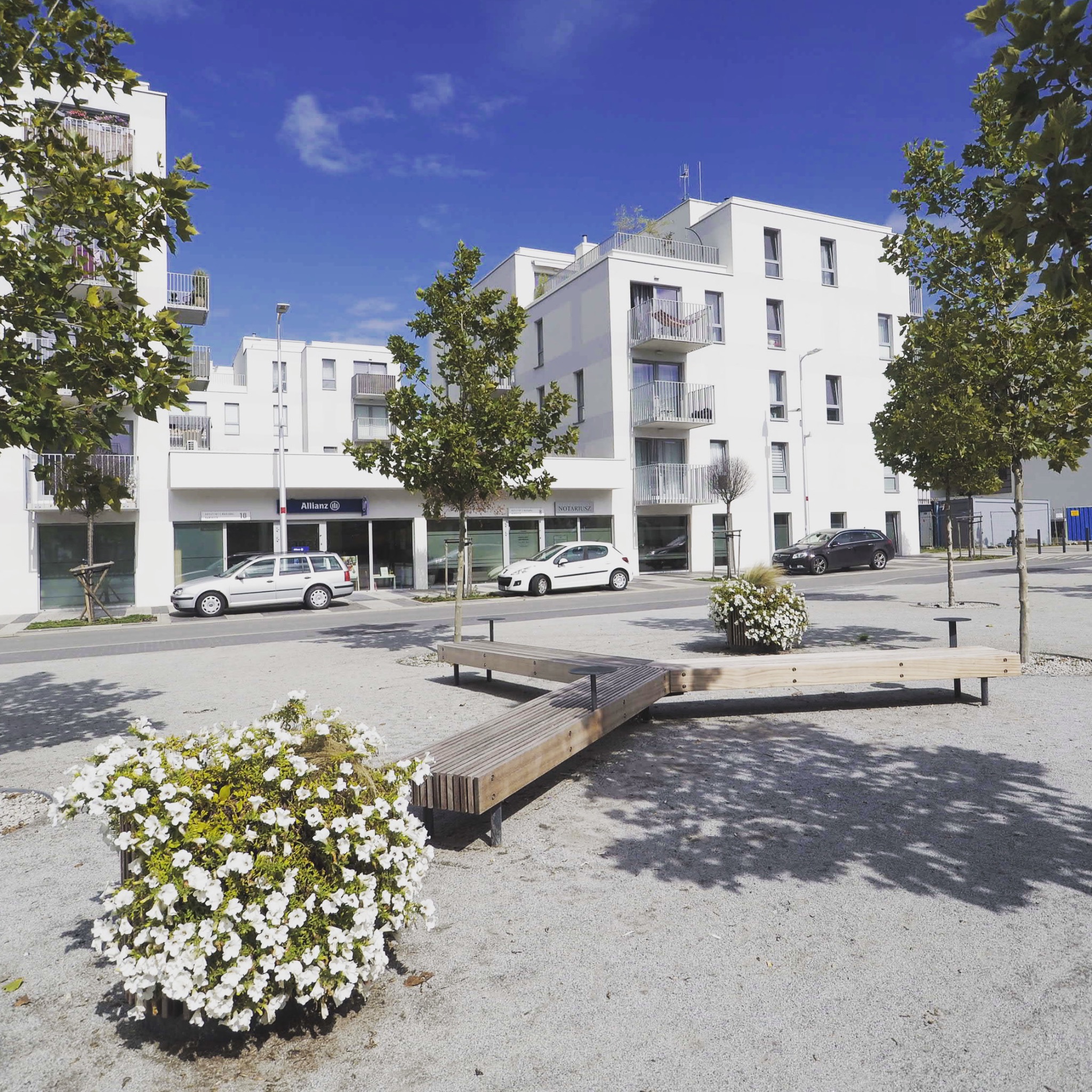 Nowoczesny biały budynek usługowy z balkonami, widok z zewnątrz na plac z ławkami i kwiatami, błękitne niebo.