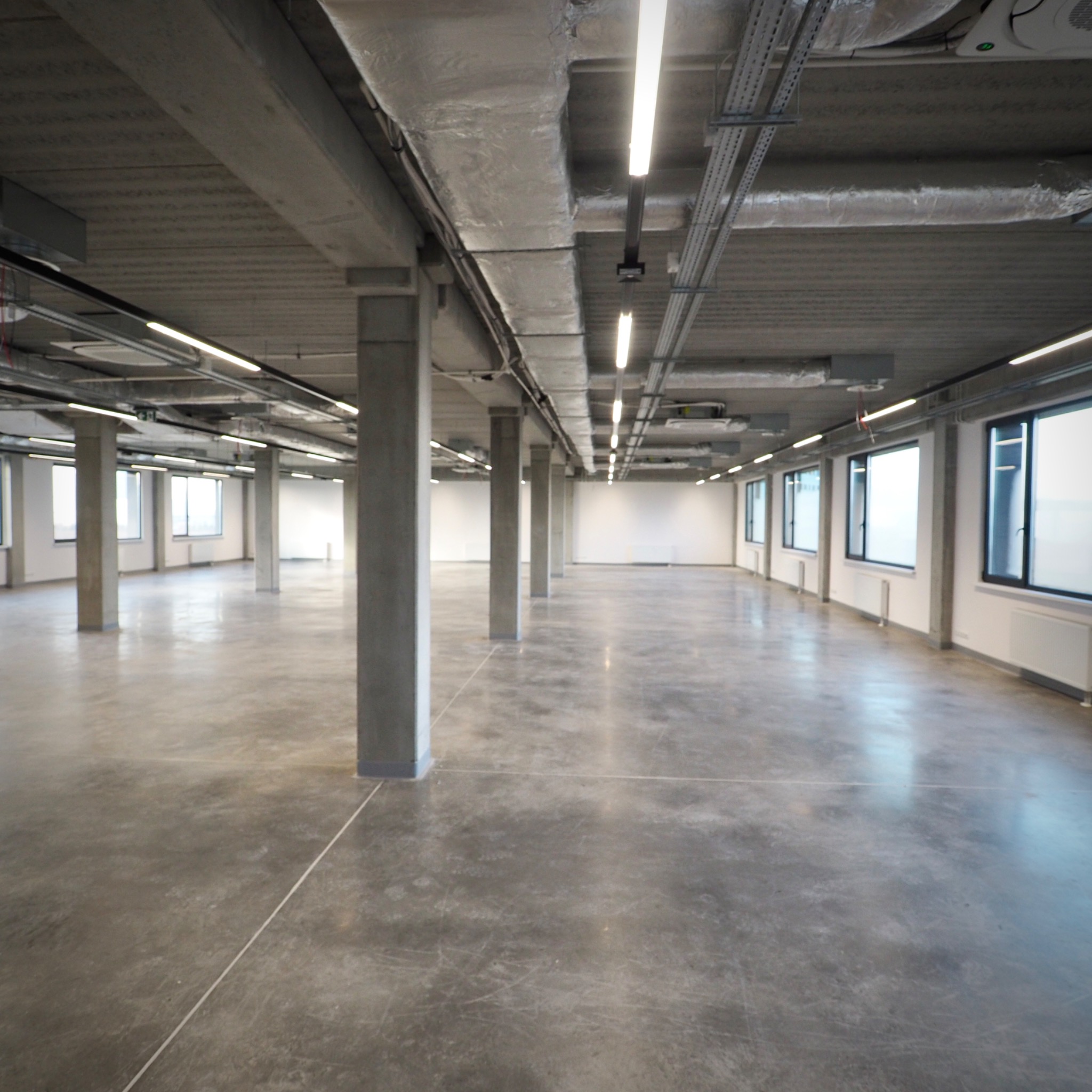 Przestronne, puste wnętrze biurowe z betonową podłogą, widoczne kolumny nośne, okna wzdłuż ściany i instalacje pod sufitem, przygotowane do aranżacji.