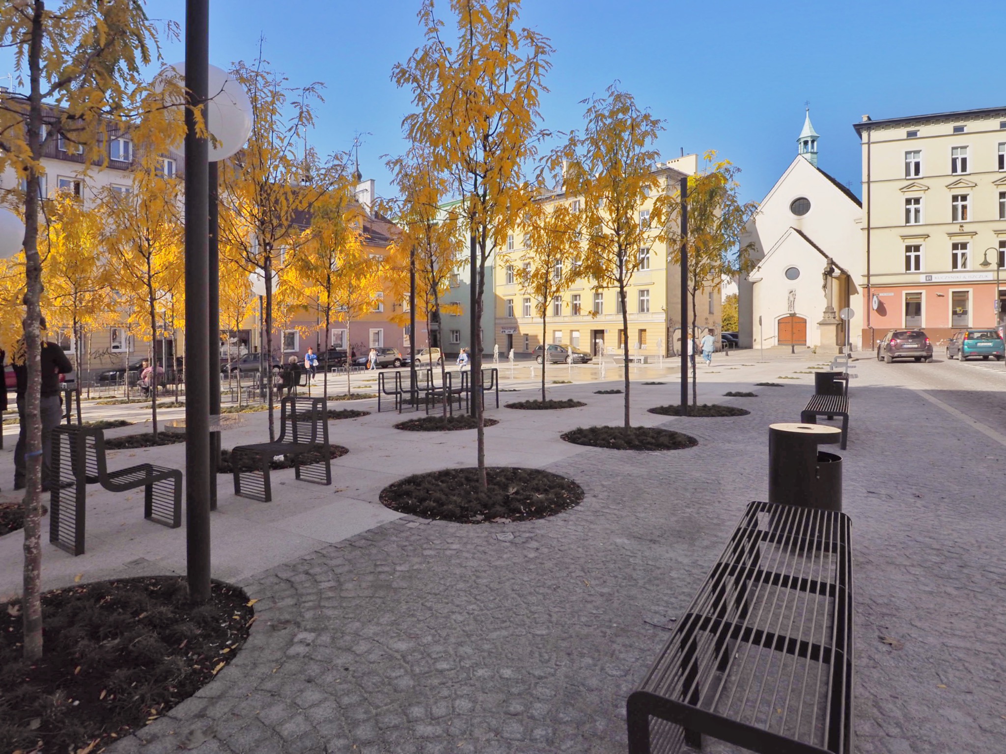 Miejska przestrzeń publiczna z rzędem drzew o żółtych liściach, nowoczesnymi ławkami z metalu i brukowaną nawierzchnią, w tle budynki mieszkalne i kościół