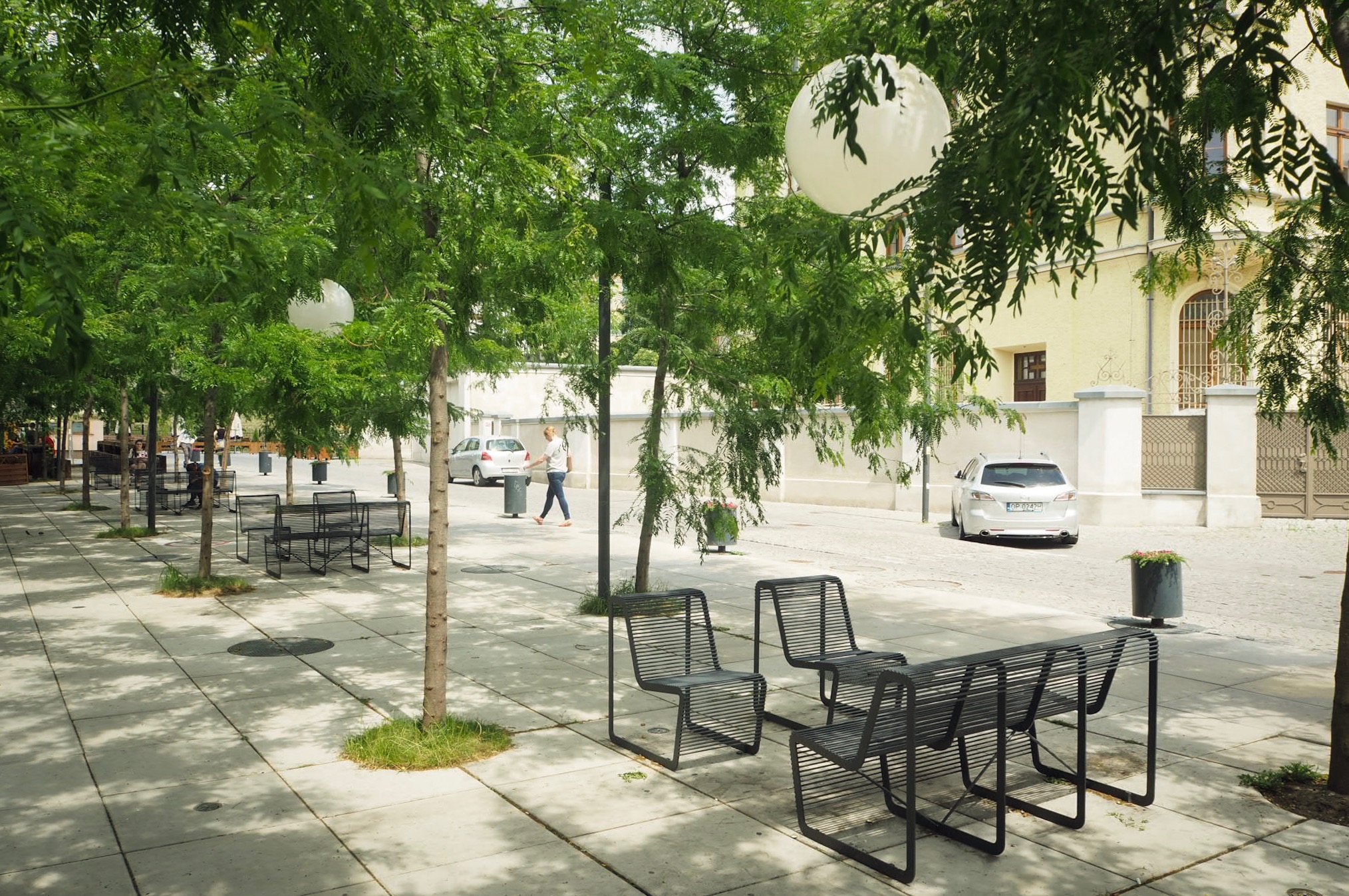 Ulica miejska z nowoczesnymi metalowymi ławkami i krzesłami, drzewami z kulistymi lampionami, w tle budynek z jasną elewacją i zaparkowany samochód.