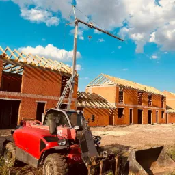 Wznoszenie szeregu budynk&oacute;w z czerwonej cegły, widoczna konstrukcja drewniana dachu oraz dźwig budowlany na tle błękitnego nieba.