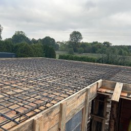 Bud-Bud Usługi ogolnobudowlane - Zbrojenie płyty fundamentowej z drewnianym szalunkiem, widok na las w tle, pochmurne niebo.