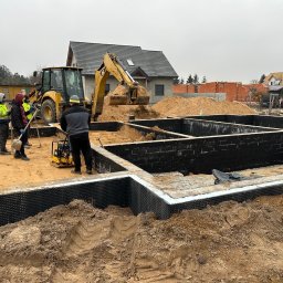 Bud-Bud Usługi ogolnobudowlane - Budowa fundamentów domu: ekipa budowlana przy użyciu koparki zasypuje piaskiem zabezpieczone folią hydroizolacyjną ściany fundamentowe pomalowane na czarno.