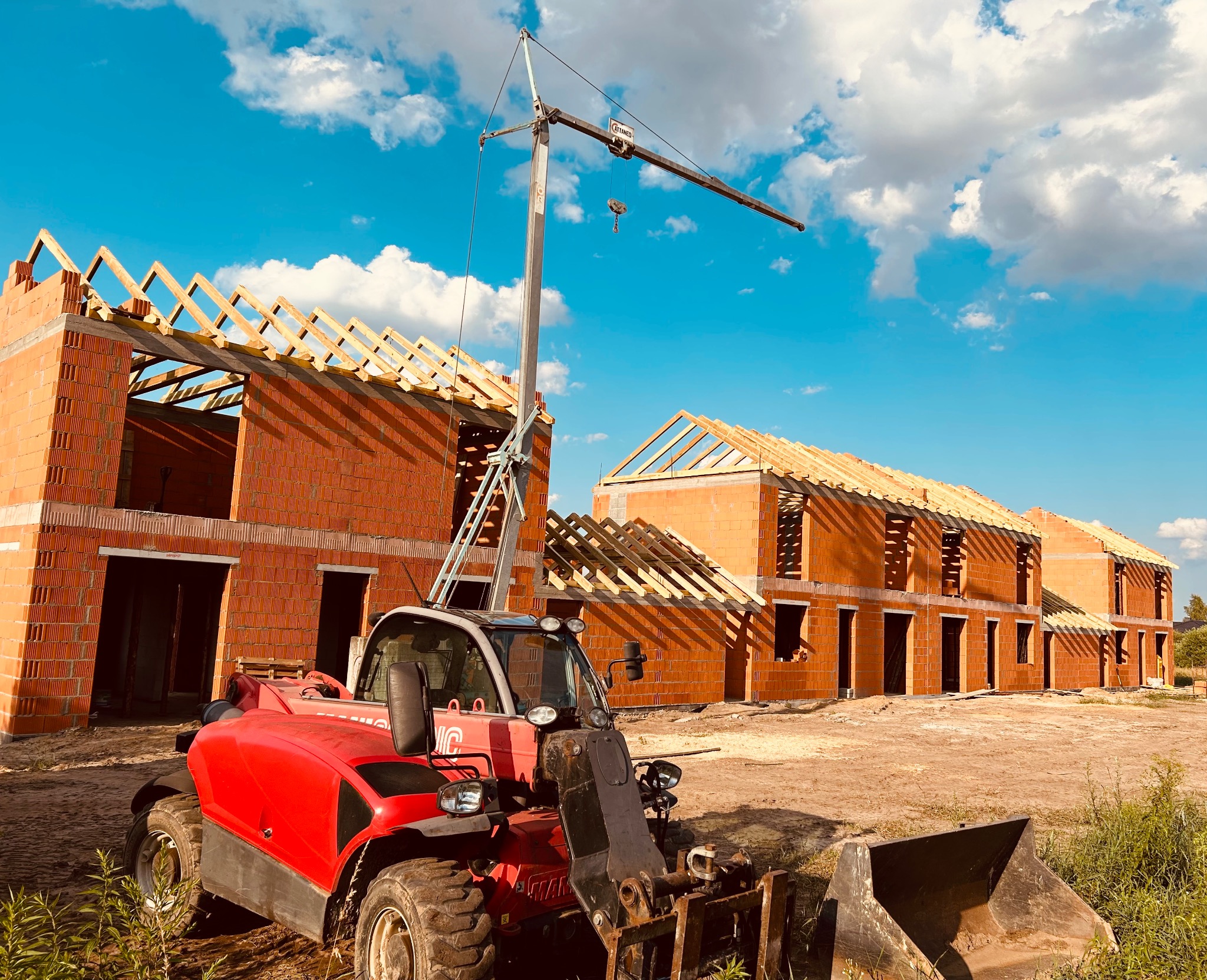 Wznoszenie szeregu budynków z czerwonej cegły, widoczna konstrukcja drewniana dachu oraz dźwig budowlany na tle błękitnego nieba.