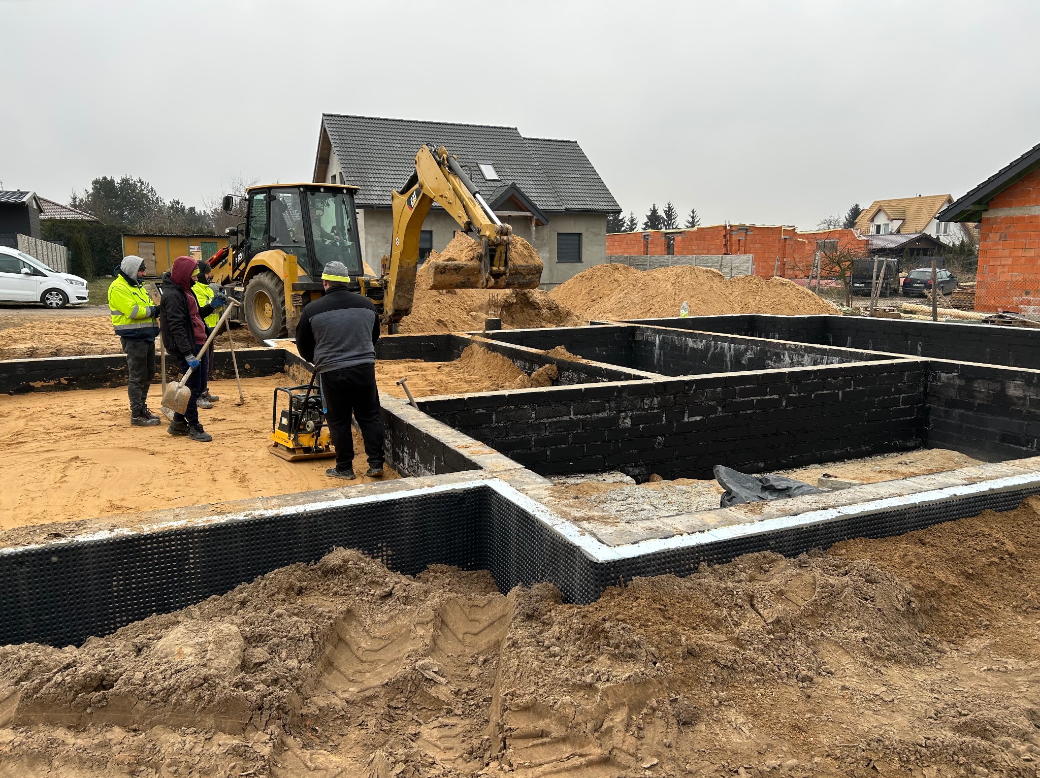 Budowa fundamentów domu: ekipa budowlana przy użyciu koparki zasypuje piaskiem zabezpieczone folią hydroizolacyjną ściany fundamentowe pomalowane na czarno.