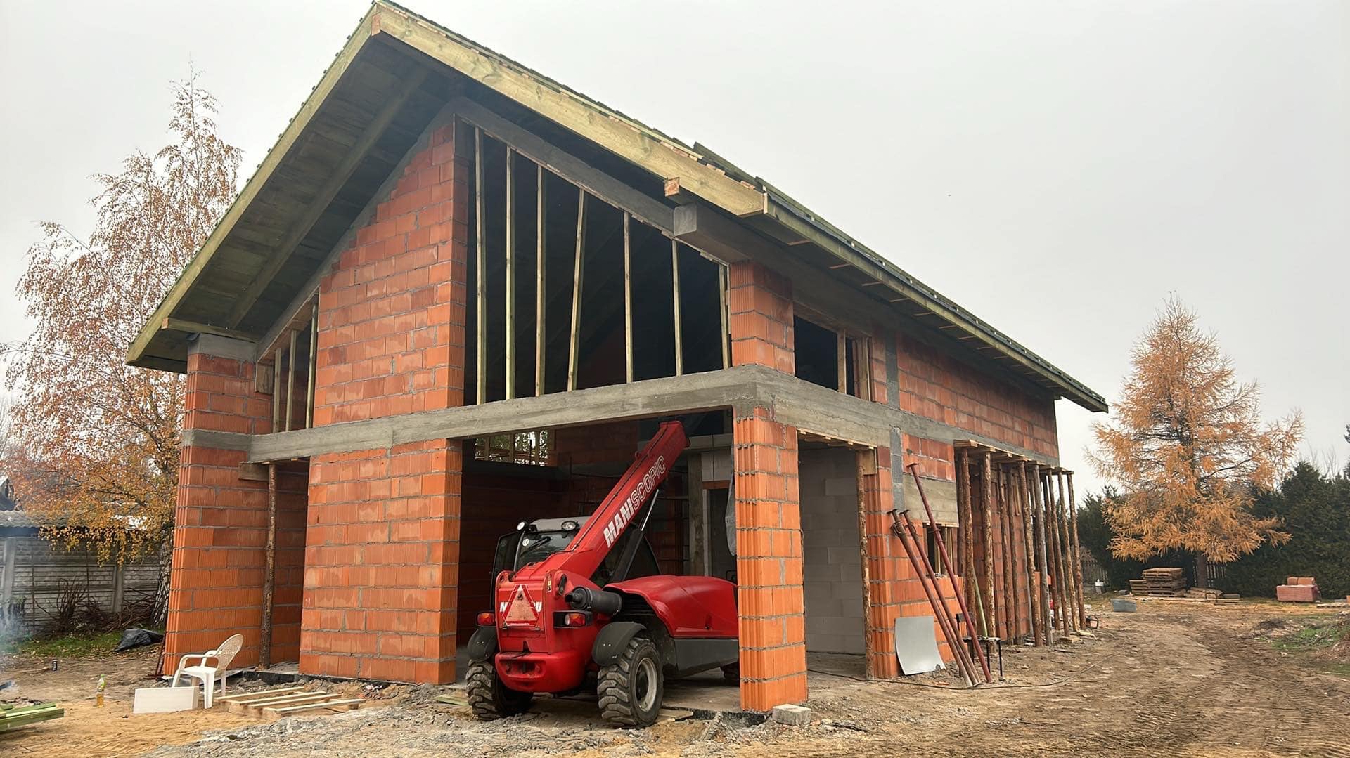 Budowa domu w stanie surowym otwartym z czerwonej cegły z widoczną więźbą dachową i podnośnikiem teleskopowym Maniscopic na placu budowy w pochmurny dzień.