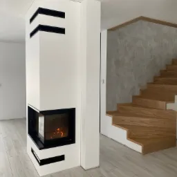 Nowoczesny kominek narożny w obudowie z białego gipsu z czarnymi wnękami, płomienie widoczne za szybą, drewniane schody w tle, podłoga wyłożona panelami w odcieniach szarości.