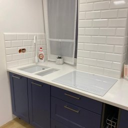 Renovation4You - Nowoczesna kuchnia z granatowymi szafkami, białym blatem, płytą indukcyjną i białą, cegiełkową glazurą, fragment niezakrytej instalacji elektrycznej nad zlewem.