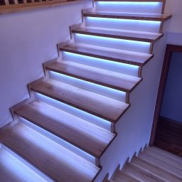 Renovation4You - Drewniane schody z podświetleniem LED wbudowanym pod każdym stopniem, oświetlające klatkę schodową z drewnianą balustradą na górze.