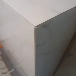 Narożnik obudowy z płytek imitujących marmur, widoczne precyzyjne fugi i listwa wykończeniowa, fragment podłogi w tle.