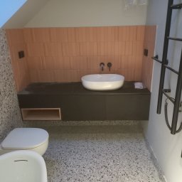 Renovation4You - Nowoczesna łazienka z umywalką nablatową, brązowymi płytkami ułożonymi pionowo, szarą szafką pod umywalką, toaletą w tle i grzejnikiem drabinkowym po prawej stronie na tle podłogi lastryko.