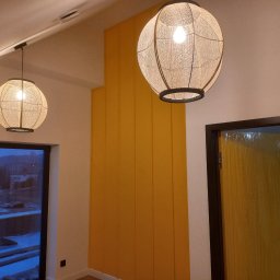 Renovation4You - Nowoczesne oświetlenie wnętrza: dwa okrągłe abażury z plecionki zawieszone na tle żółtej ściany panelowej i okna balkonowego z widokiem na zimowy krajobraz.