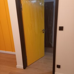 Renovation4You - Żółte drzwi z czarną ościeżnicą, zabezpieczone folią ochronną, otwarte w nowym mieszkaniu z panelami podłogowymi i pomarańczową ścianą w tle.