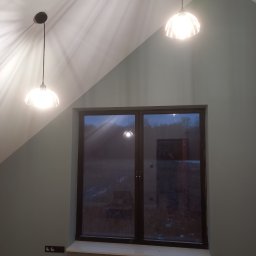 Renovation4You - Nowoczesne wnętrze z oknem w ciemnej ramie, dwiema wiszącymi lampami z kloszami w stylu retro i detektorem dymu na skośnym suficie, ściany pomalowane na stonowany odcień zieleni.