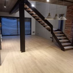 Renovation4You - Nowoczesne wnętrze z jasną drewnianą podłogą, schodami z metalową konstrukcją i drewnianymi stopniami zabezpieczonymi folią, betonowym sufitem i trzema wiszącymi lampami z białymi kloszami
