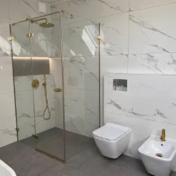 Nowoczesna łazienka z kabiną prysznicową typu walk-in z elementami w kolorze złotym, marmurowe płytki na ścianach, podwieszana toaleta i bidet, szara podłoga.