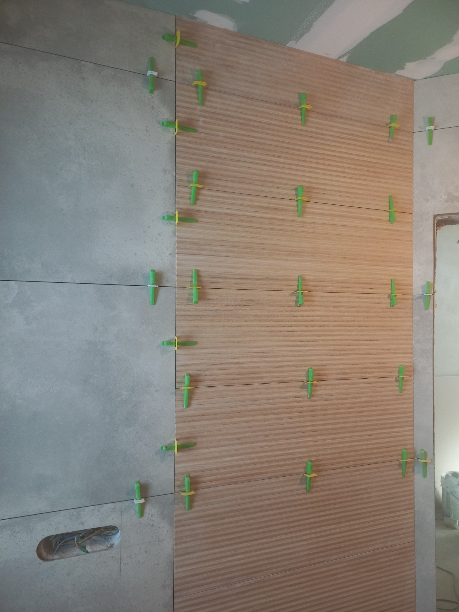 Ściana wyłożona częściowo szarymi płytkami, a częściowo płytkami imitującymi drewno, z widocznymi krzyżykami dystansowymi i poziomowaniem laserowym, otwór w płytkach na instalację elektryczną.