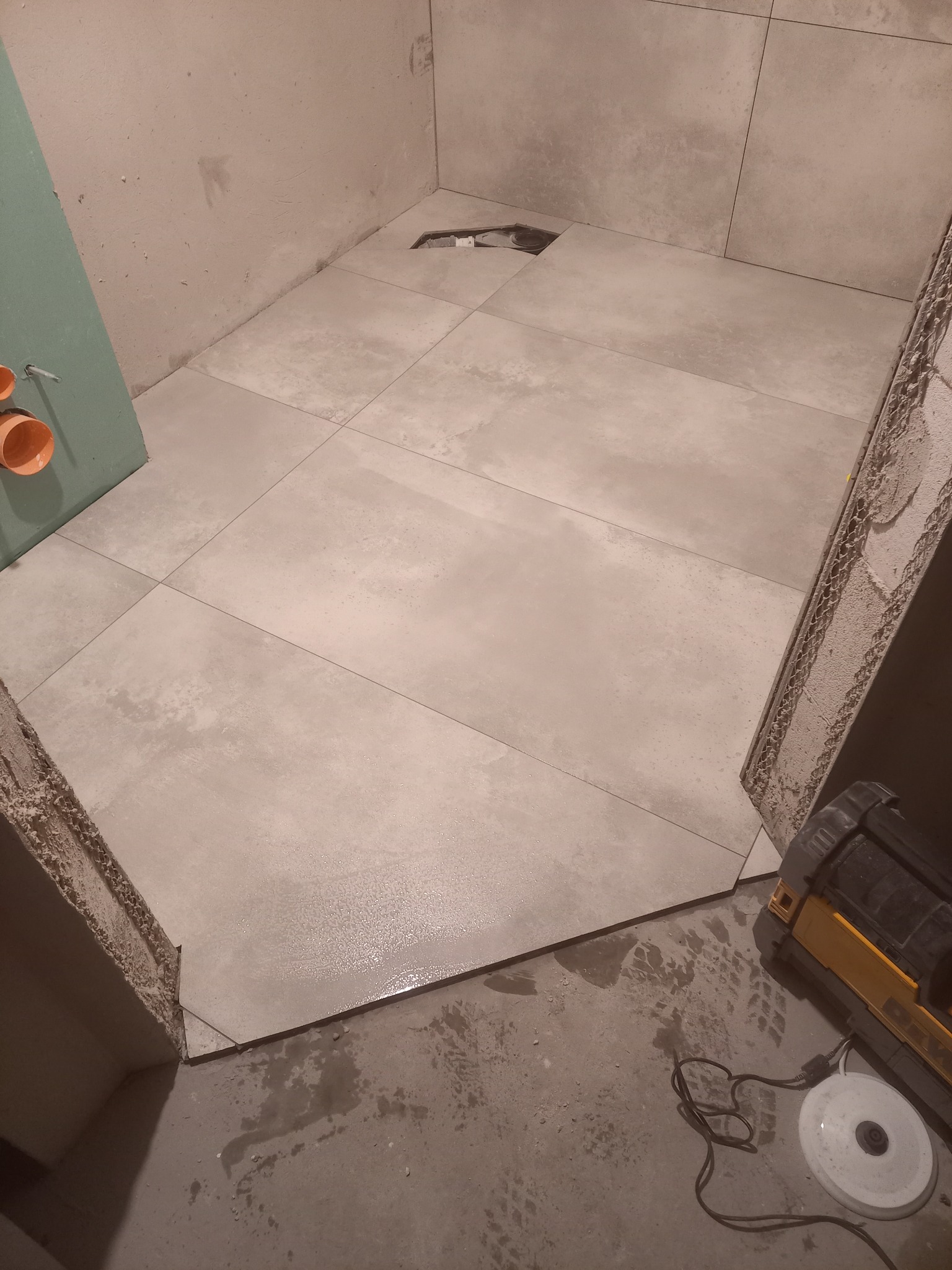 Ułożone szare płytki podłogowe w trakcie prac w łazience, widoczny odpływ liniowy, fragment ściany z instalacją hydrauliczną i narzędzia na podłodze.