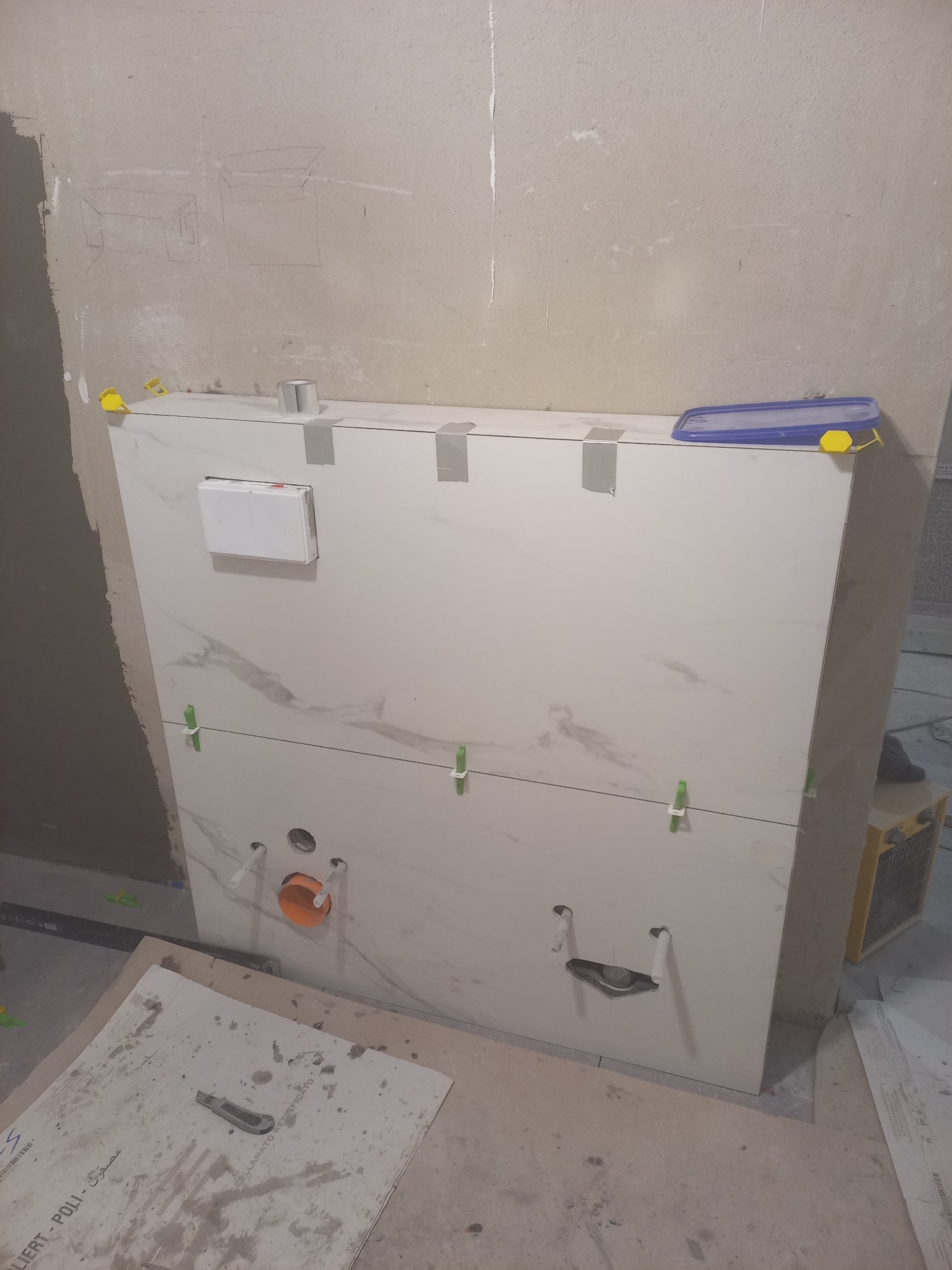 W trakcie montażu stelaża podtynkowego WC z płytami imitującymi marmur, widoczne zielone krzyżyki dystansowe, taśma maskująca i otwory na przyłącza, na podłodze płyta z nożem do cięcia.