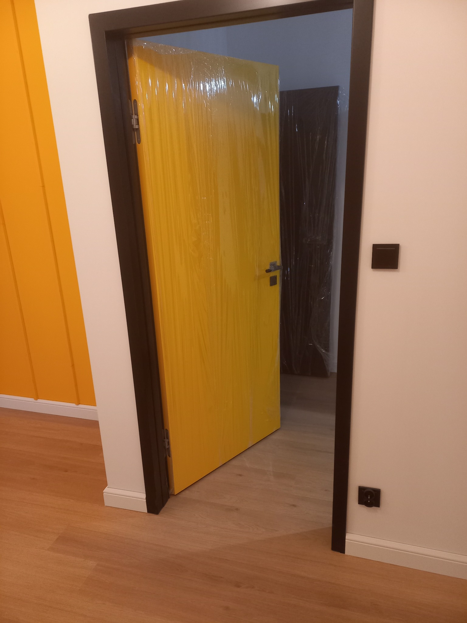 Żółte drzwi z czarną ościeżnicą, zabezpieczone folią ochronną, otwarte w nowym mieszkaniu z panelami podłogowymi i pomarańczową ścianą w tle.