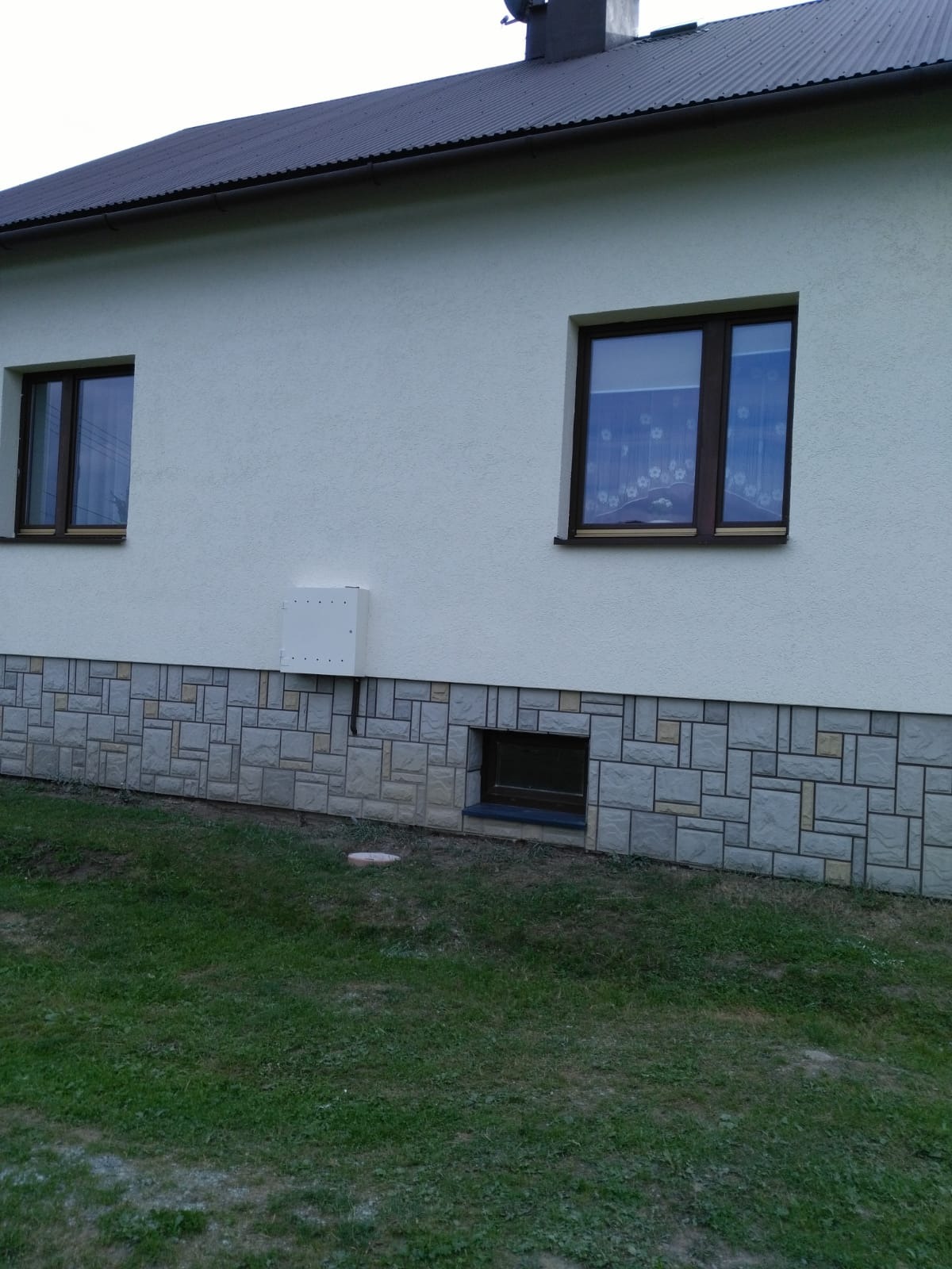 Elewacja domu z oknami, wykończona kamienną okładziną w dolnej części, z widoczną skrzynką elektryczną i oknem piwnicznym.