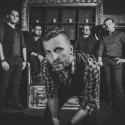 Czarno-białe portretowe ujęcie pięciu mężczyzn w eleganckich strojach, jeden z nich w pierwszym planie w koszuli w kratę i kamizelce, pochylony do przodu, pozostałych w tle, siedzących...