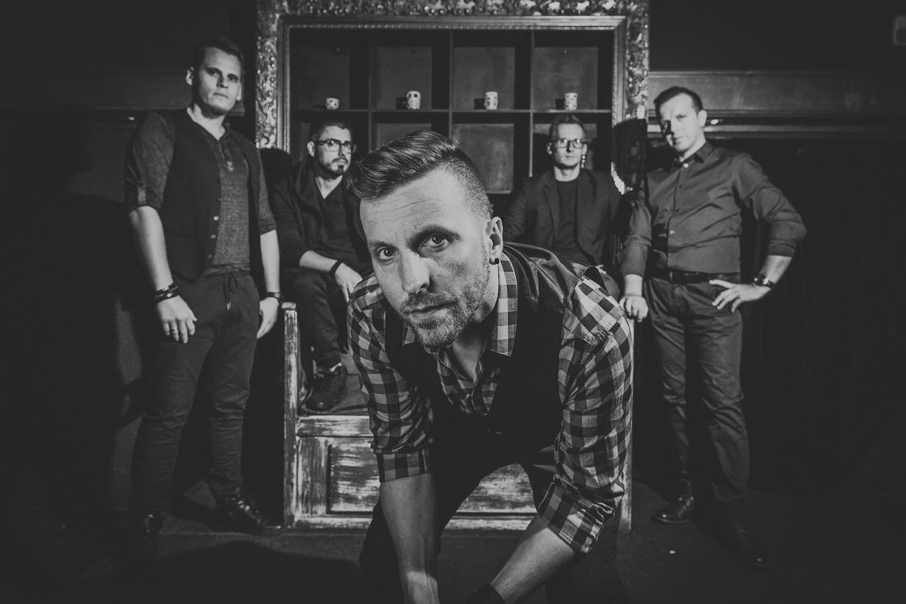 Czarno-białe portretowe ujęcie pięciu mężczyzn w eleganckich strojach, jeden z nich w pierwszym planie w koszuli w kratę i kamizelce, pochylony do przodu, pozostałych w tle, siedzących...