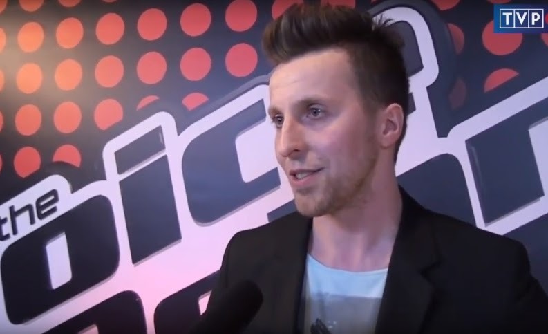 Młody mężczyzna w czarnej marynarce udziela wywiadu na tle logo programu The Voice i logotypu TVP.