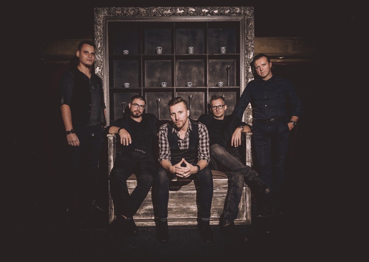 Portret studyjny pięciu mężczyzn w ciemnych ubraniach, siedzących i stojących na tle dekoracyjnej ramy z półkami, w stonowanej kolorystyce.