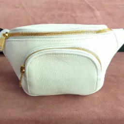 Biała skórzana nerka z złotymi zamkami błyskawicznymi na brązowym tle, widoczny czarny pasek z klamrą.