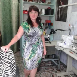 Krawcowa w sukience w motywy zwierzęce, stojąca w pracowni z maszynami do szycia i regałem z akcesoriami krawieckimi.