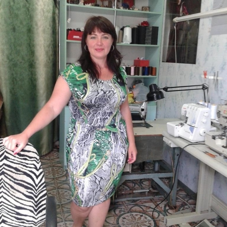 Krawcowa w sukience w motywy zwierzęce, stojąca w pracowni z maszynami do szycia i regałem z akcesoriami krawieckimi.