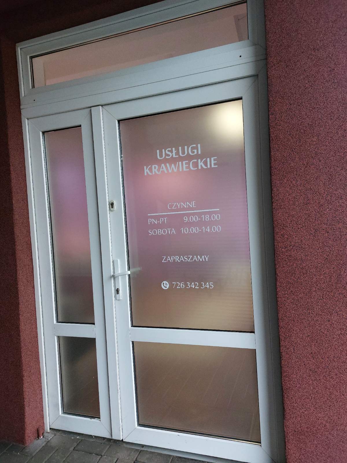 Białe drzwi wejściowe do punktu usługowego z naklejonym napisem 'Usługi Krawieckie' oraz informacją o godzinach otwarcia i numerem telefonu.