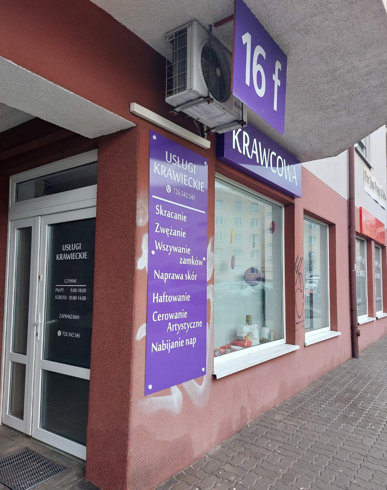 Fasada budynku z szyldem 'Usługi Krawieckie' oferującego skracanie, zwężanie, wszywanie zamków, naprawę skór, haftowanie, cerowanie artystyczne i nabijanie nap, widoczny numer telefonu i godziny...