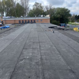 Domidach - Widok z góry na płaski dach pokryty papą, z ekipą dekarzy pracującą w tle przy krawędzi budynku, narzędzia i materiały budowlane rozłożone na powierzchni dachu. W tle widoczne drzewa i budynki.