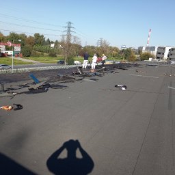Domidach - Ekipa dekarzy w trakcie demontażu starego pokrycia bitumicznego na dachu płaskim budynku przemysłowego. W tle widoczne kominy przemysłowe i budynki mieszkalne.