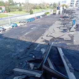 Domidach - Demontaż starego, zniszczonego pokrycia dachowego płaskiego dachu budynku, widoczne fragmenty papy, ekipa w białych kombinezonach w oddali, linia zaparkowanych samochodów wzdłuż krawędzi dachu.