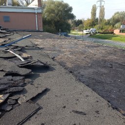 Domidach - Demontaż starego pokrycia dachowego z papy, widoczne fragmenty odspojonej izolacji, przygotowanie pod nowe krycie, w tle budynek i słupy wysokiego napięcia.