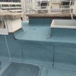 Niebieska papa na płaskim dachu z kominem wentylacyjnym i świetlikiem dachowym na tle zabudowy miejskiej.