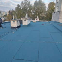 Domidach - Niebieska płaska powierzchnia dachu pokryta papą termozgrzewalną, z widocznymi kominami wentylacyjnymi i sylwetką pracownika w oddali, na tle bloków mieszkalnych i drzew.