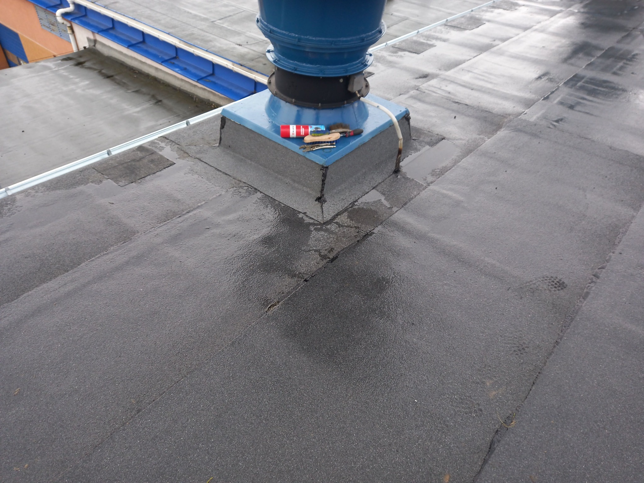 Płaski, mokry dach pokryty papą z niebieską podstawą komina wentylacyjnego i narzędziami dekarskimi (pędzel, szczypce, puszka kleju) leżącymi obok niego.