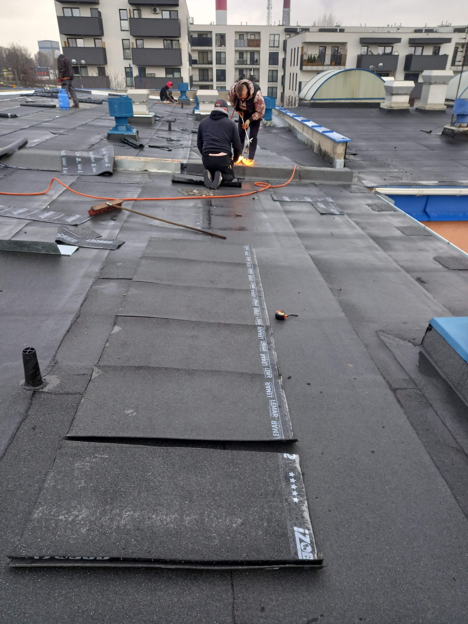 Ekipa dekarzy pracuje na płaskim dachu bloku, używając palnika do łączenia bitumicznej membrany dachowej, widoczne narzędzia i częściowo położona izolacja.
