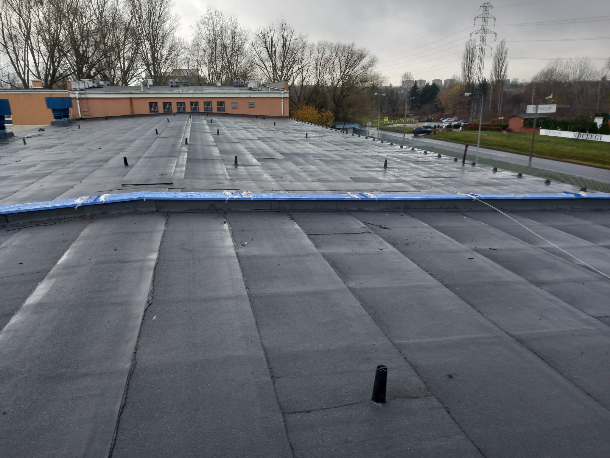 Widok z góry na płaski dach pokryty papą termozgrzewalną z widocznymi kominkami wentylacyjnymi, zabezpieczony niebieską folią na krawędzi, z budynkiem w tle.