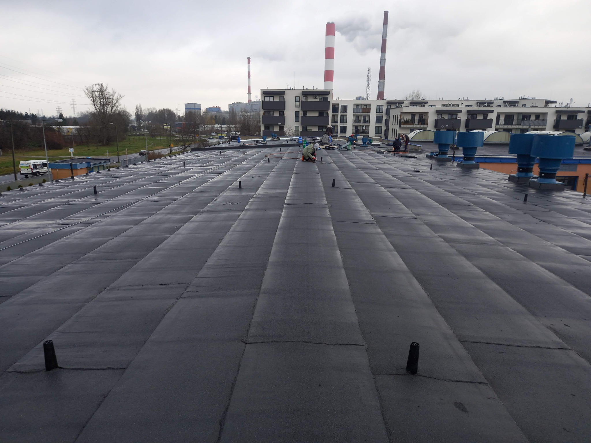 Ekipa dekarzy pracuje na płaskim dachu pokrytym czarnym materiałem bitumicznym, w tle kominy elektrowni i budynki mieszkalne.