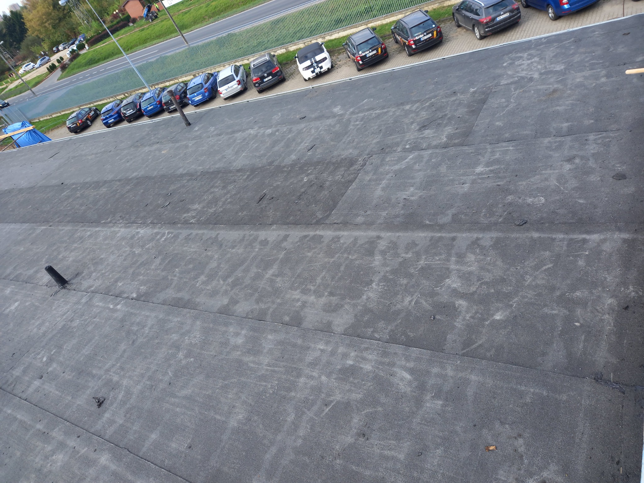 Widok z góry na płaski, czarny dach pokryty papą, z widocznymi łączeniami i kominkiem wentylacyjnym; w tle rząd zaparkowanych samochodów i zieleń.